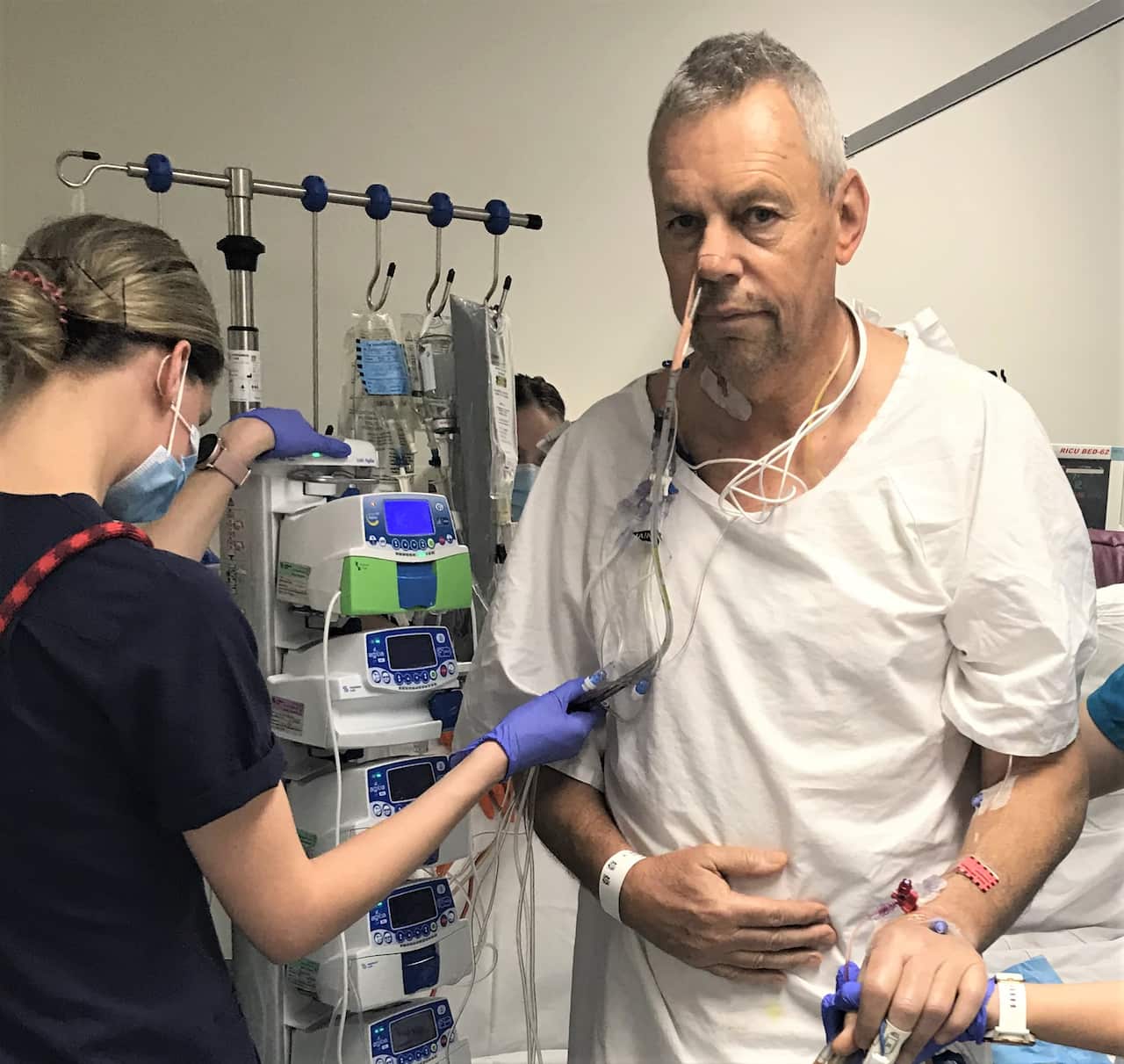 A man standing in a white gown in hospital. He has wires attached to his body. A woman standing next to him is holding some of the wires.