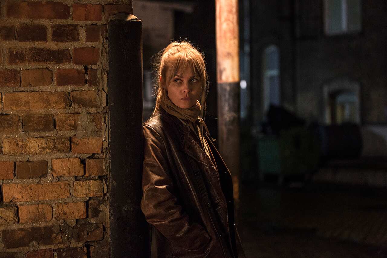 A woman stands in a dark alley, in a coat and big scarf. 