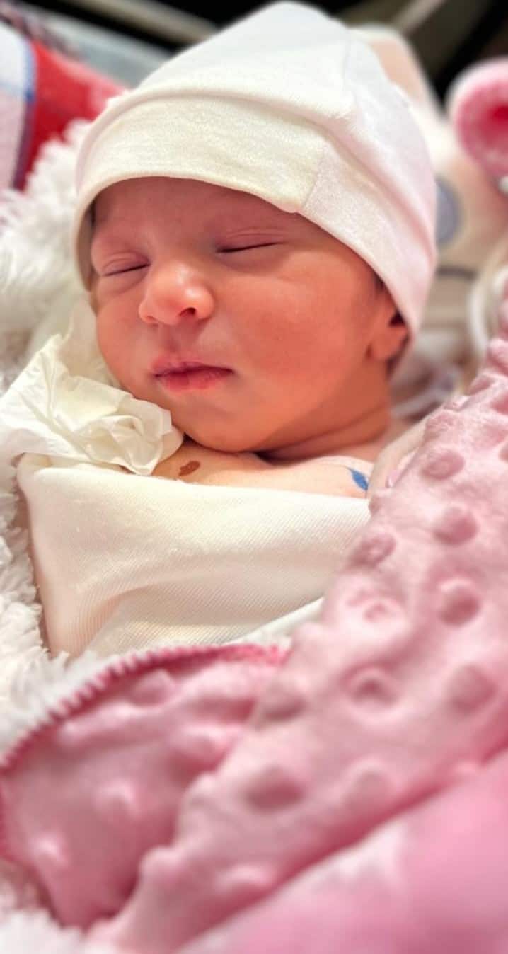 A baby girl has her eyes closed. She wears a baby and is covered with a pink blanket. 