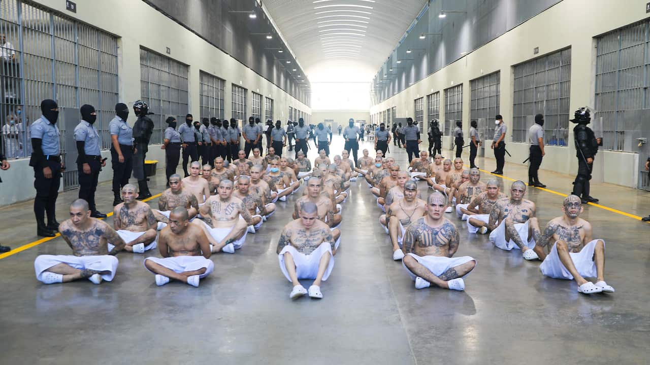 Shirtless, tattooed men with shaved heads, wearing white shorts, sitting in rows on the prison floor with their hands handcuffed behind their backs. They are surrounded by security guards wearing balaclavas.