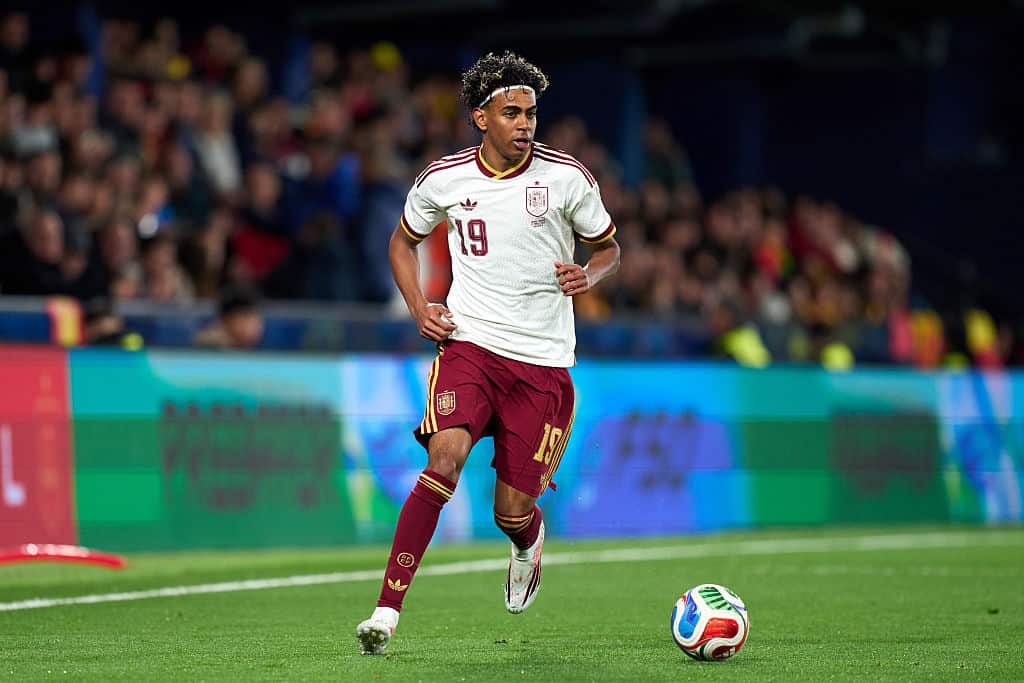 A footballer wearing a white shirt and maroon shorts running with the ball on a grass field 