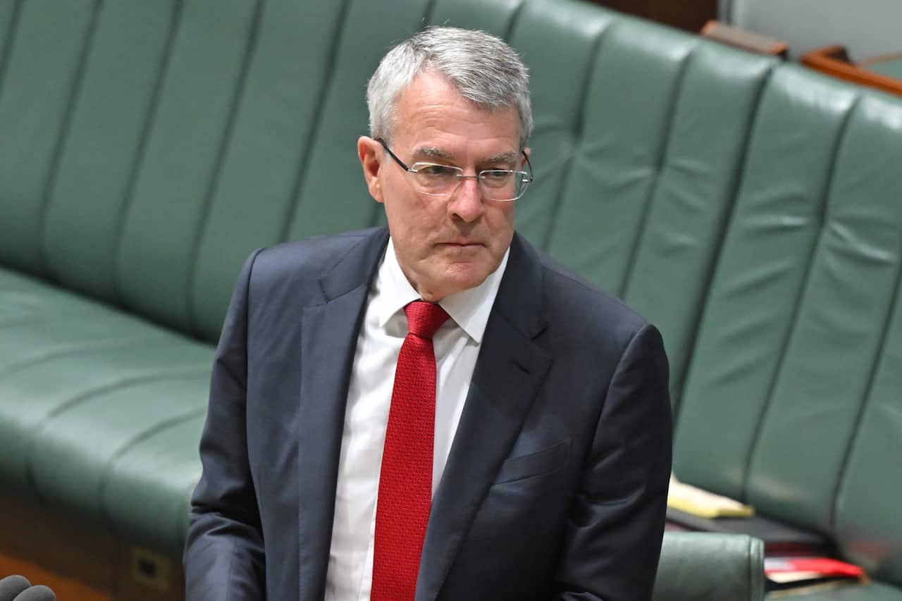 A man with grey hair, glasses, a blue blazer and red tie standing, with green seats behind him.