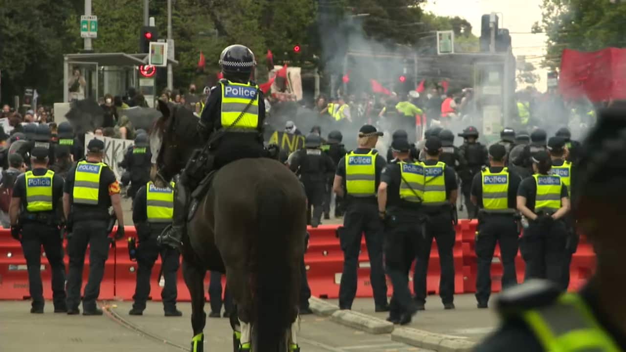 A line of Victoria Police officers in riot gear, including one on horseback, facing a cloud of smoke and a crowd of protesters.