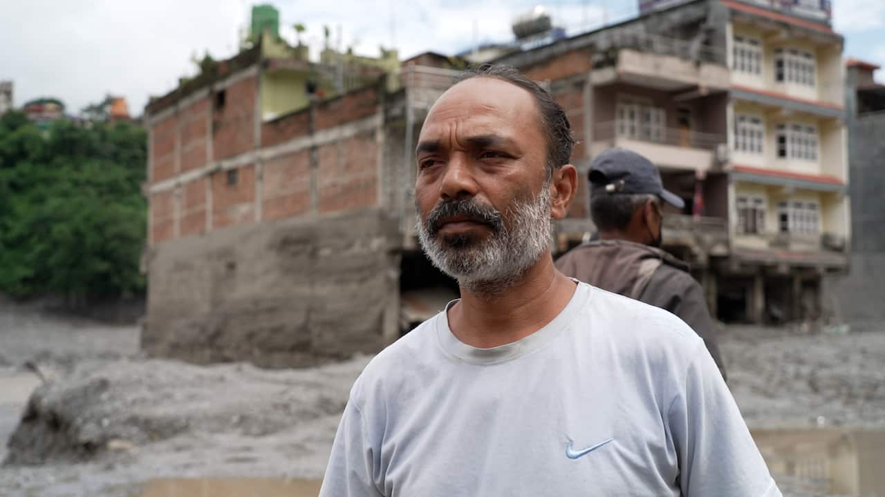 An older Nepali man with a greying beard standing in front of building that has been inundated with flood waters