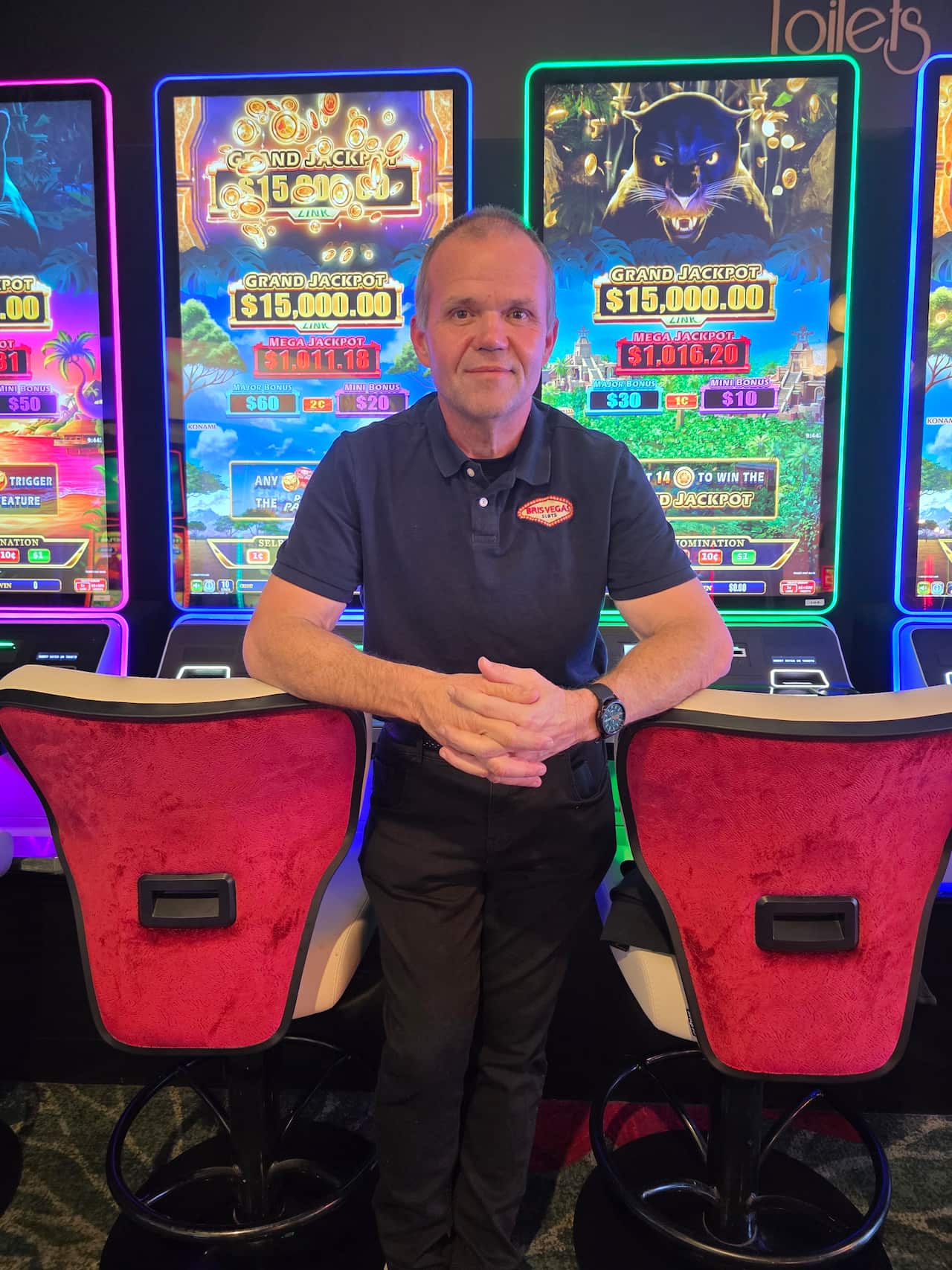 A balding man is crossing his hands wearing a black shirt with the logo brisvegas on it with several poker machines behind him