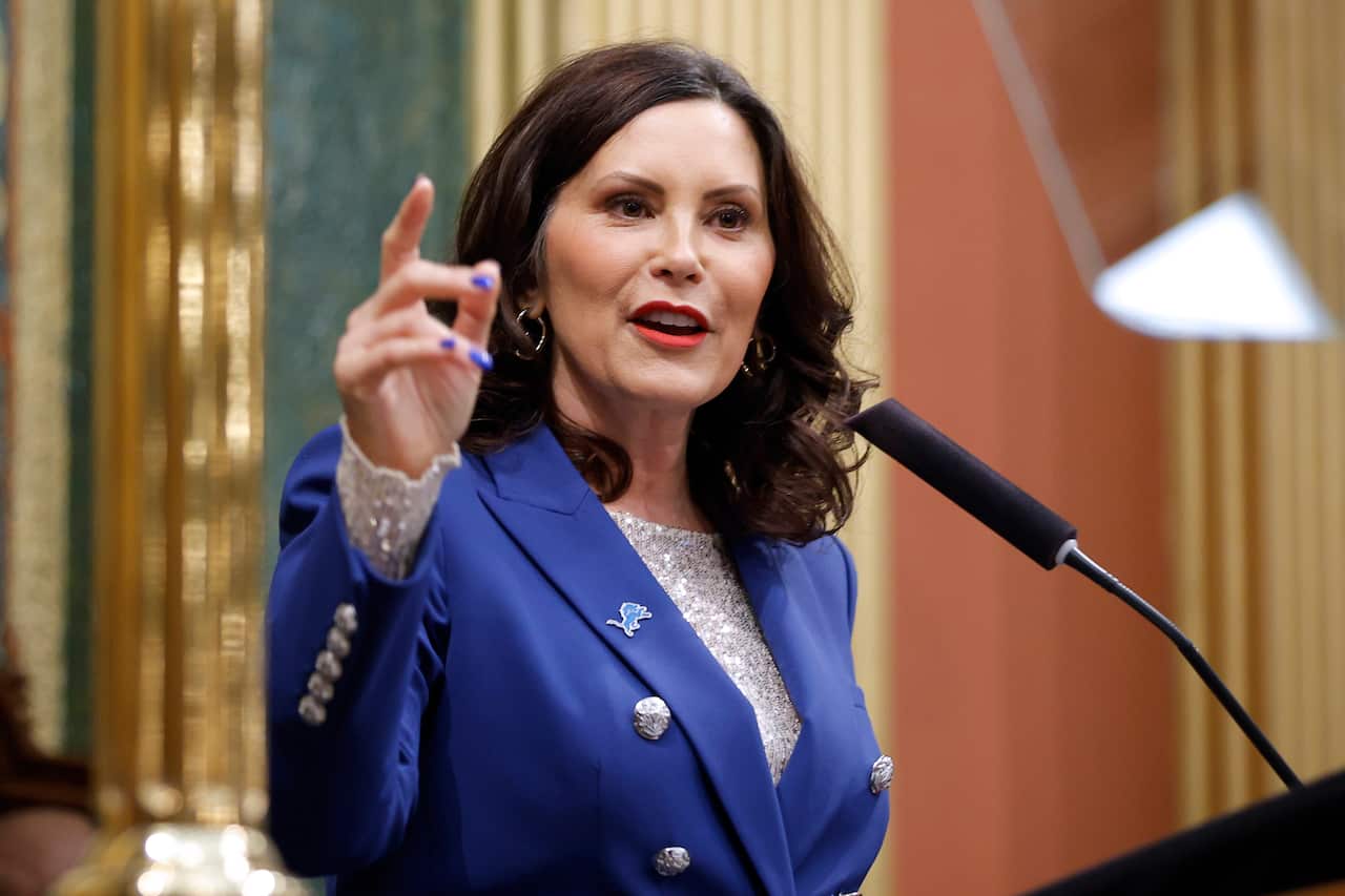 A woman with long brown hair, wearing a sparkly silver top and a blue blazer while speaking at a lecturn