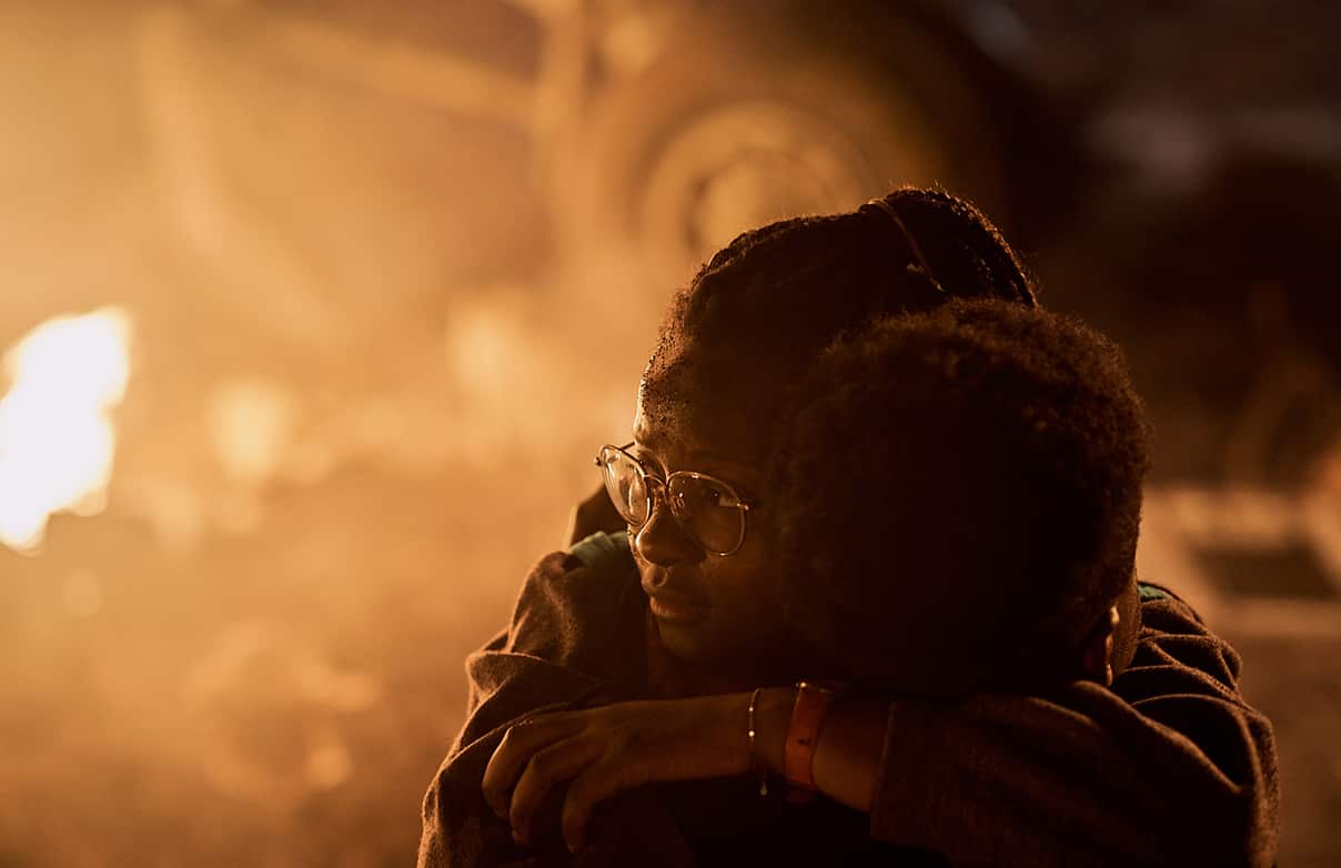 Two people huddle, looking afraid, in front of a burning background.