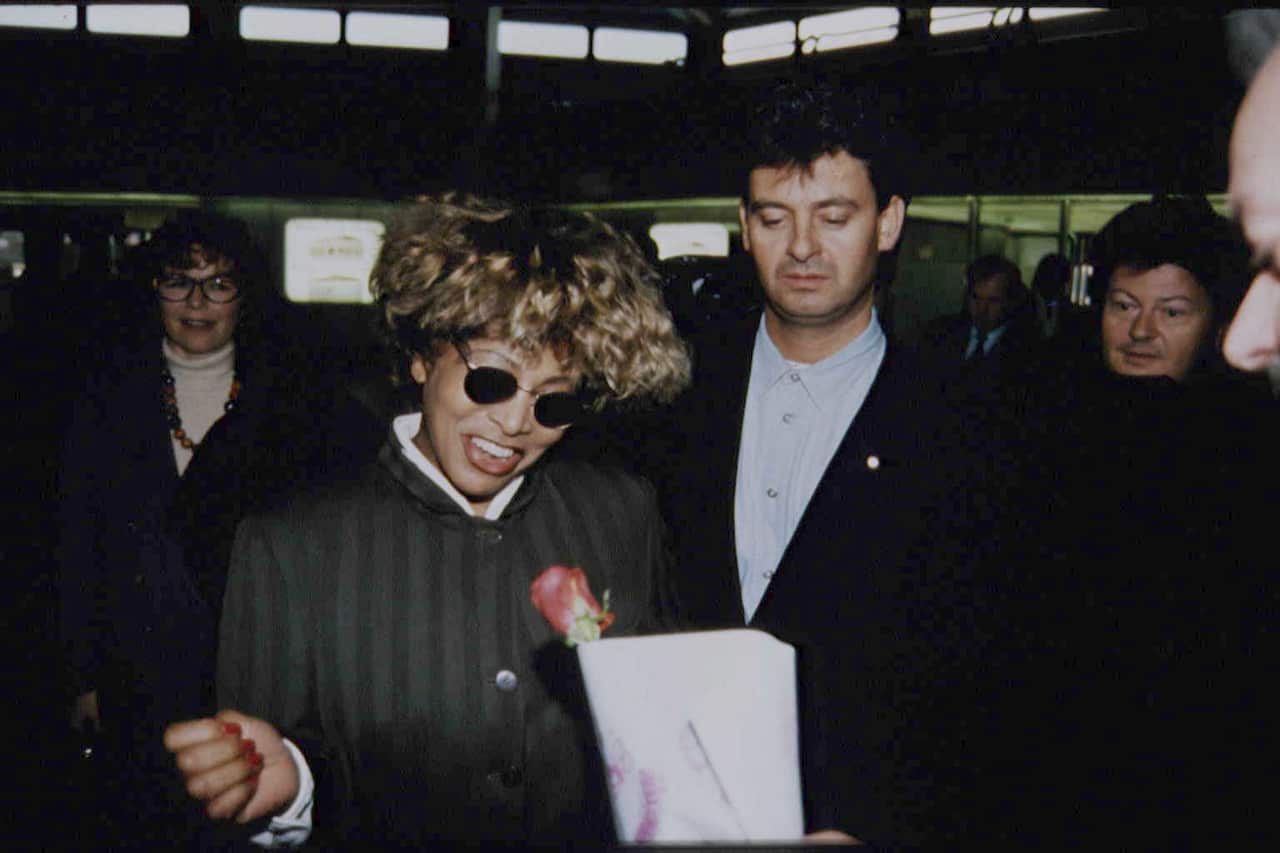 Tina Turner wears aviator sunglasses and walks through a crowd of people with Erwin Bach slightly behind her