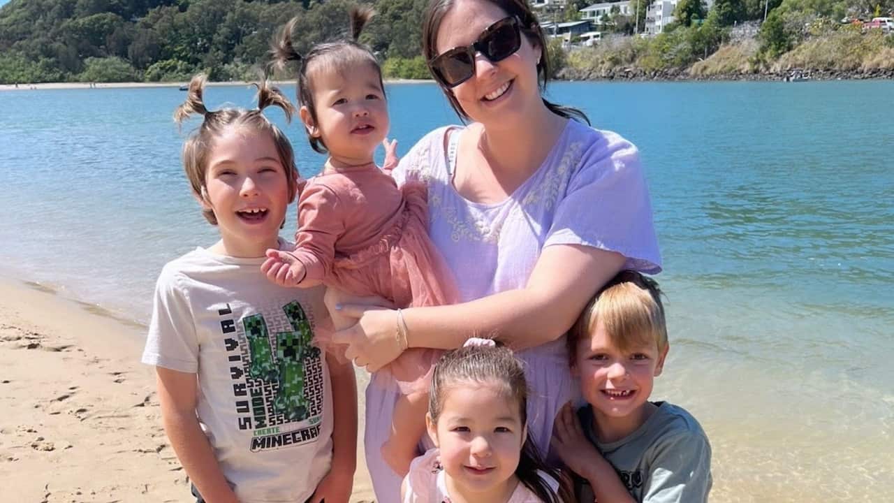 a young mum holding a young child while three other children stand close to her on a beach 
