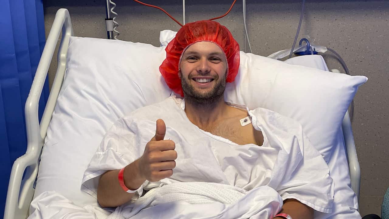 a smiling man wearing a hospital gown and cap lies in a hospital bed giving a 'thumbs-up' sign with his right hand.
