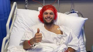 a smiling man wearing a hospital gown and cap lies in a hospital bed giving a 'thumbs-up' sign with his right hand.