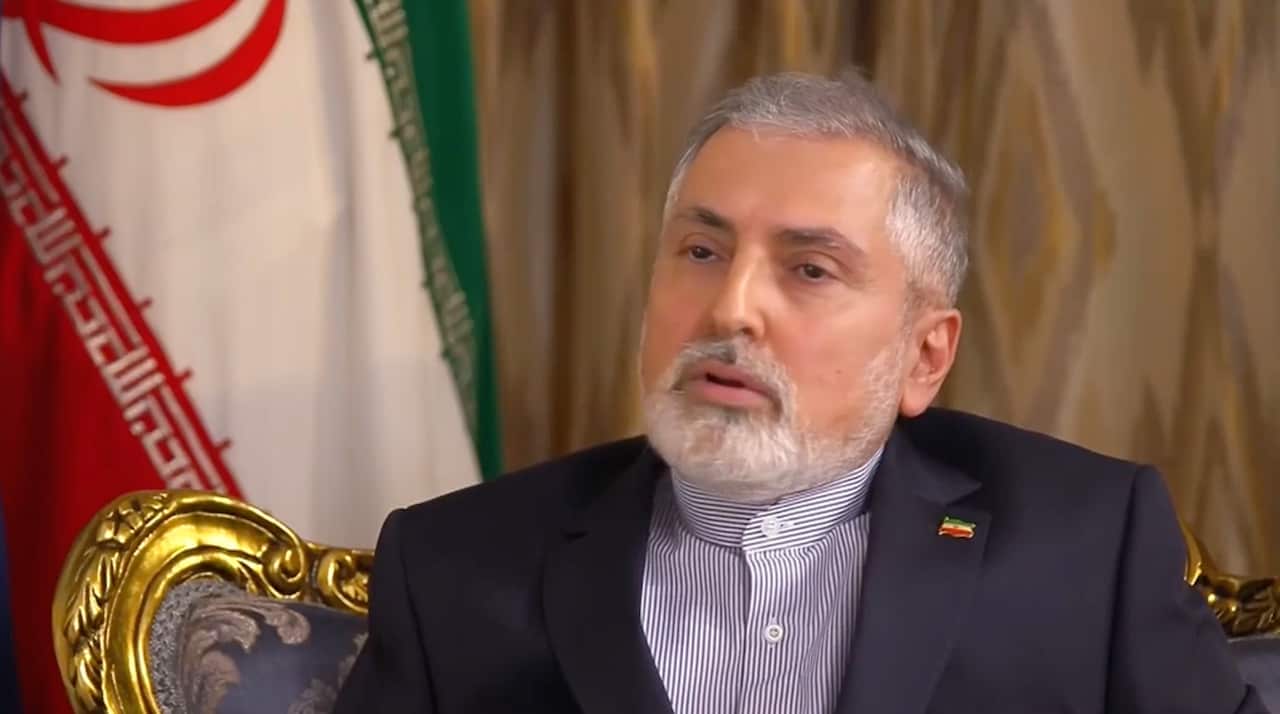 A man sitting on a chair, speaking, in front of an Iranian flag.