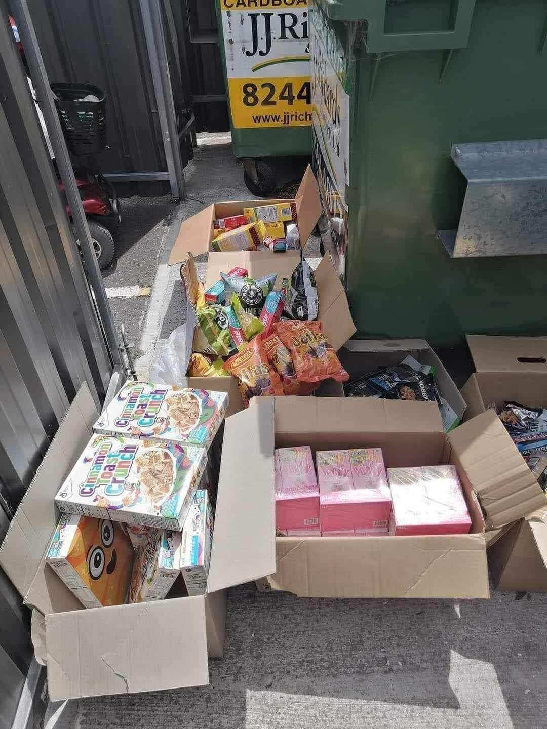 beside a bin lies several cardboard boxes packed with packaged goods including cereal and chips