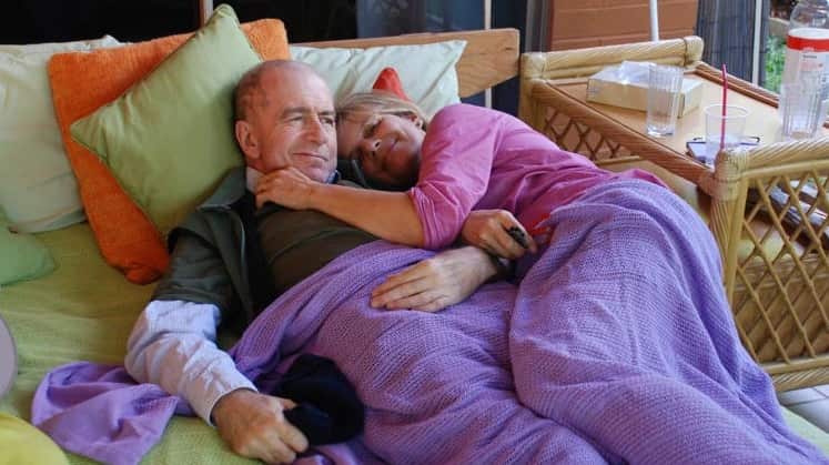A man and woman lie under a purple blanket on a bed together and embrace, fully clothed and smiling.