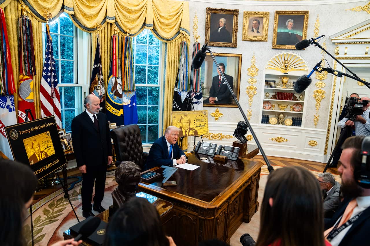 Donald Trump sitting at his desk with gold gilding on walls in the background.