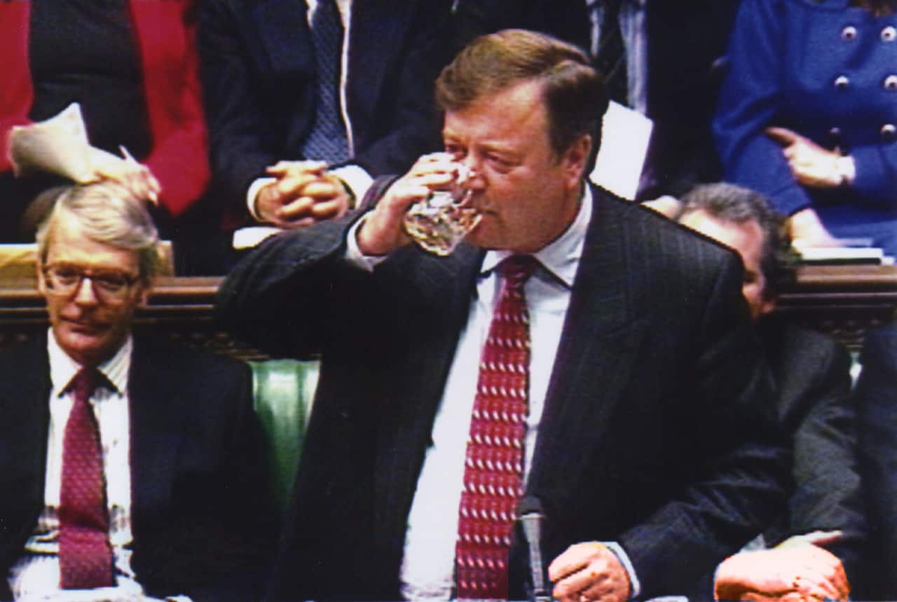 A man in a black suit and a red tie is sipping alcohol from a glass as he stands in front of a microphone inside the UK House of Commons.