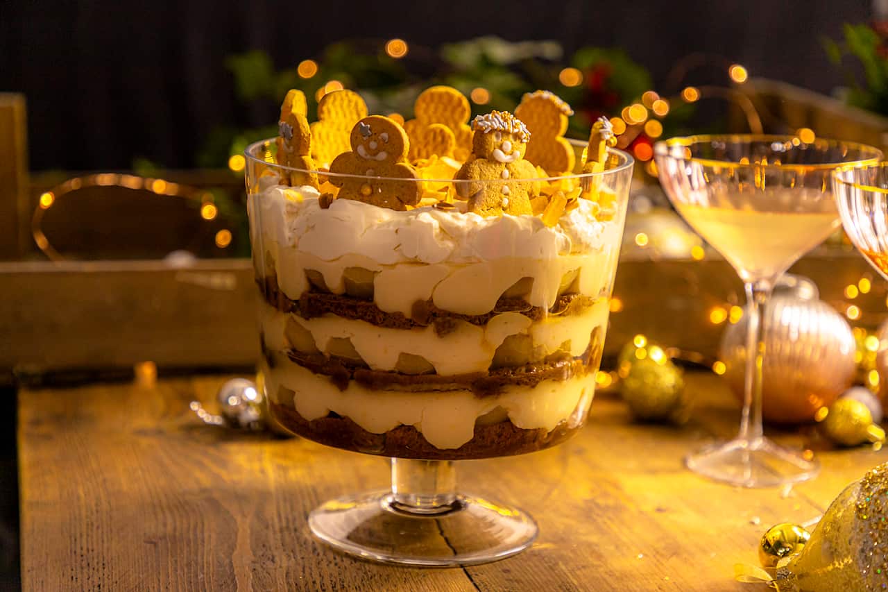 A three-layer trifle, topped with gingerbread people standing around the top edge, sits in a deep glass footed serving dish. The light is golden and festive decorations can be seen to the side.  