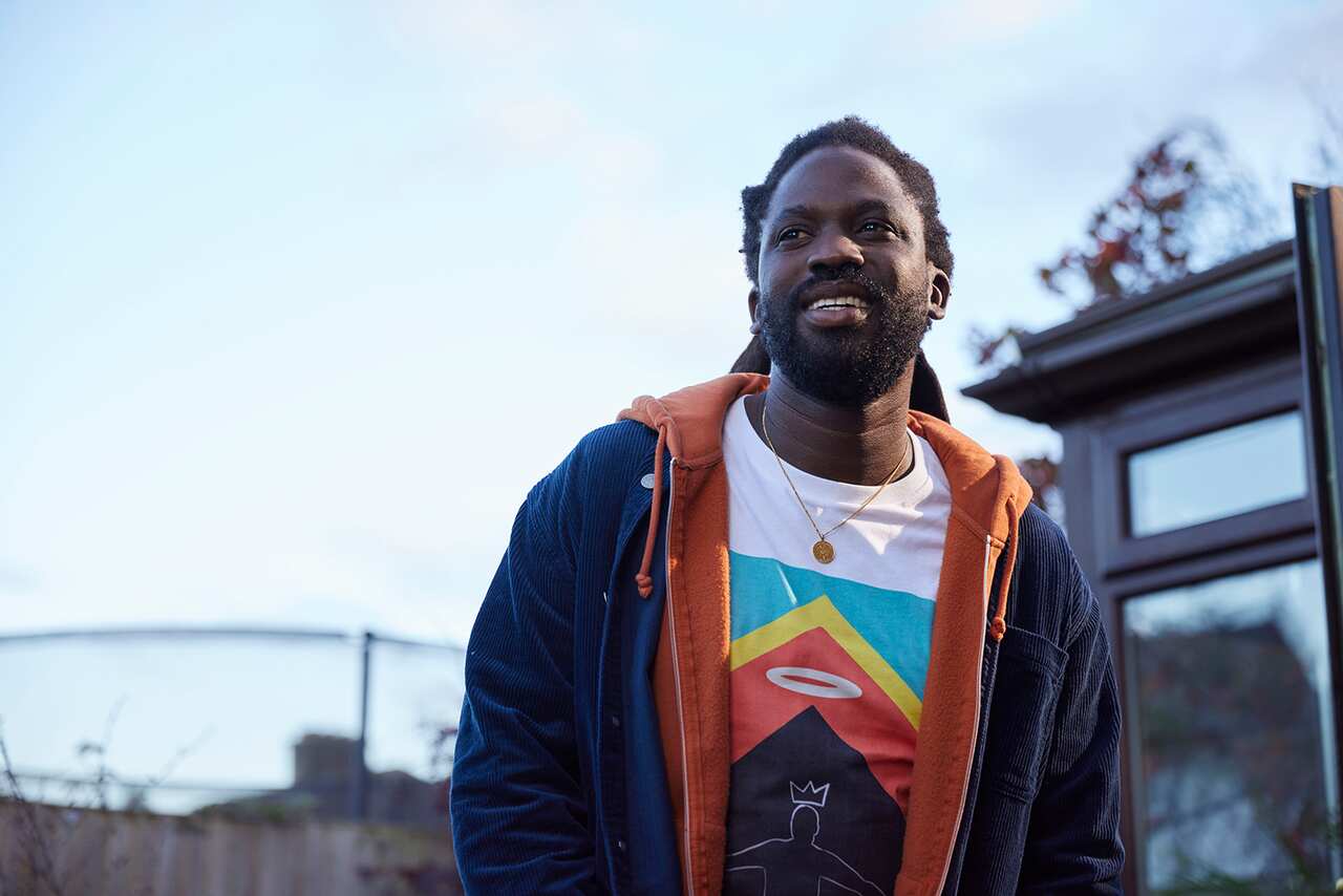 A man in a bright t-shirt and dark parka stands outdoors with a pale sky above, looking up and to the side. 
