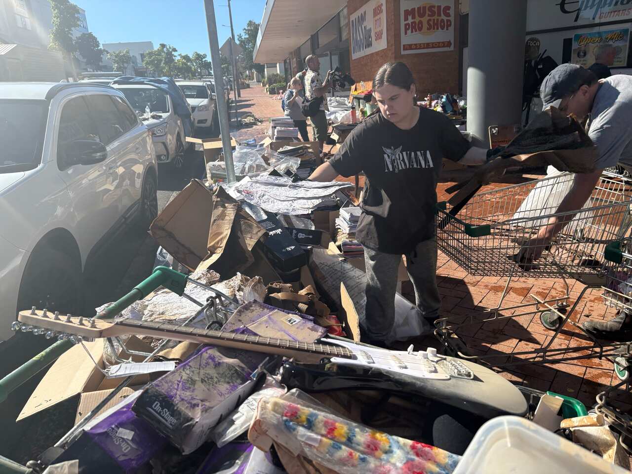 A woman in a black Nirvana T-shirt sorts damaged music shop merchandise on a storefront footpath.