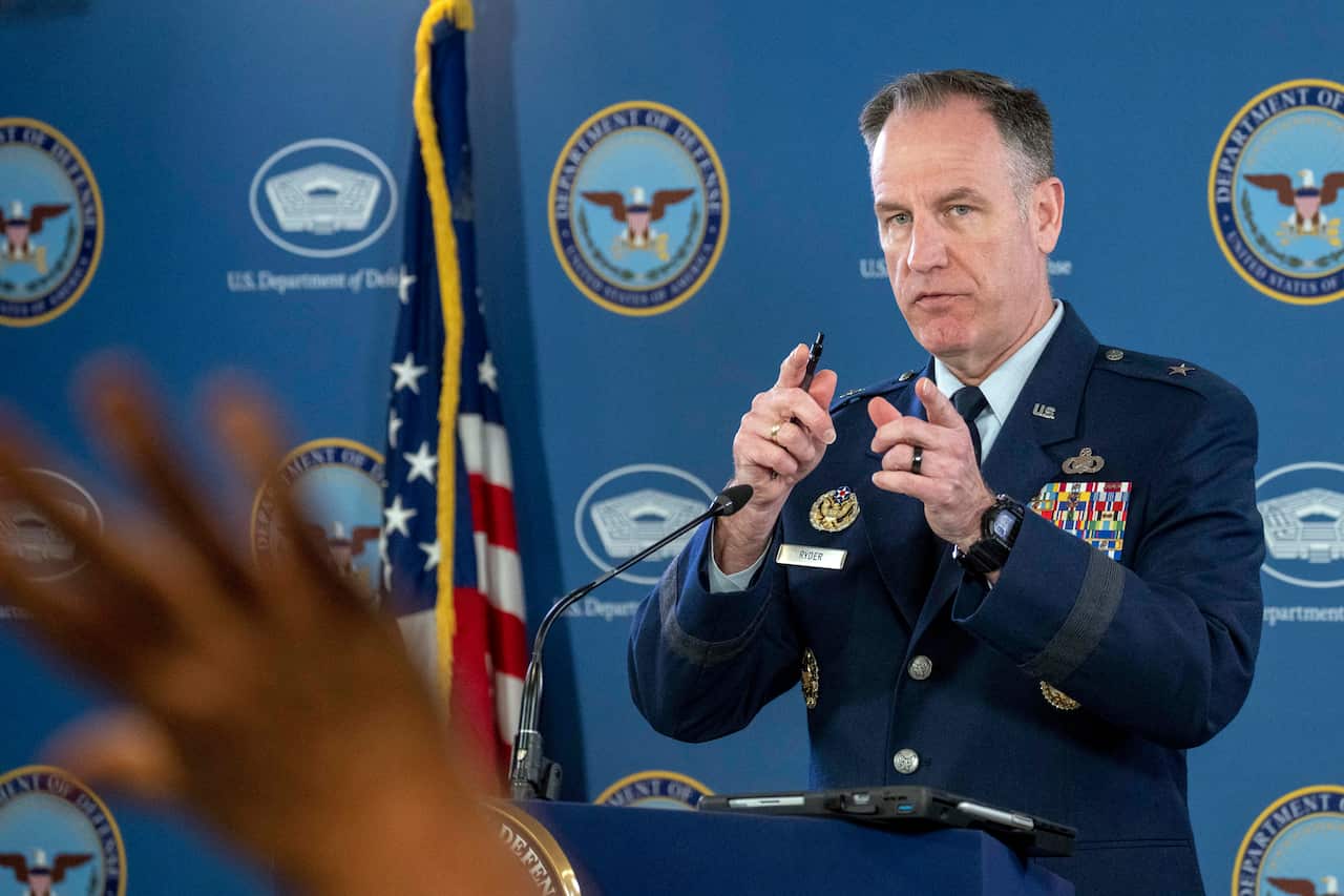 Man in a navy blue army uniform stands at the podium as he points to a raised hand waiting to ask him a question in the audience.