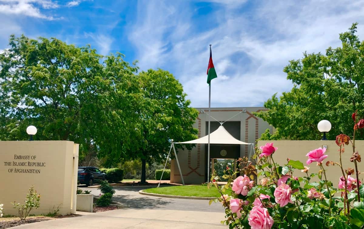 A building with a sign out the front that reads "Embassy of the Islamic Republic of Afghanistan". There are flowers and trees in the courtyard.