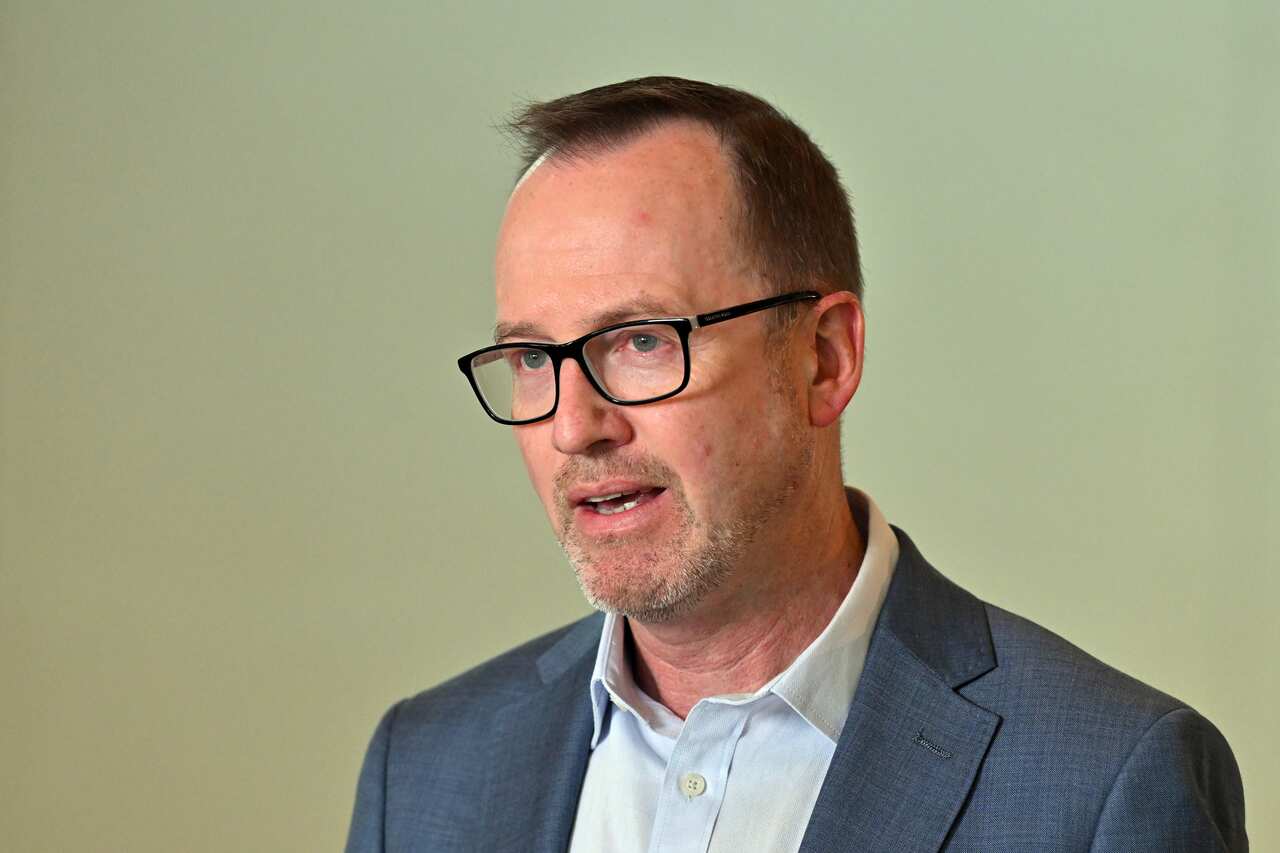 David Shoebridge wearing a blue blazer in front of a beige background.