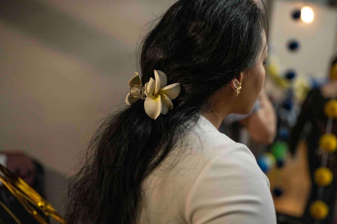 A Pasifika woman with long dark hair pulled into a ponytail with a frangipani clip is wearing a white top and facing away from the camera.