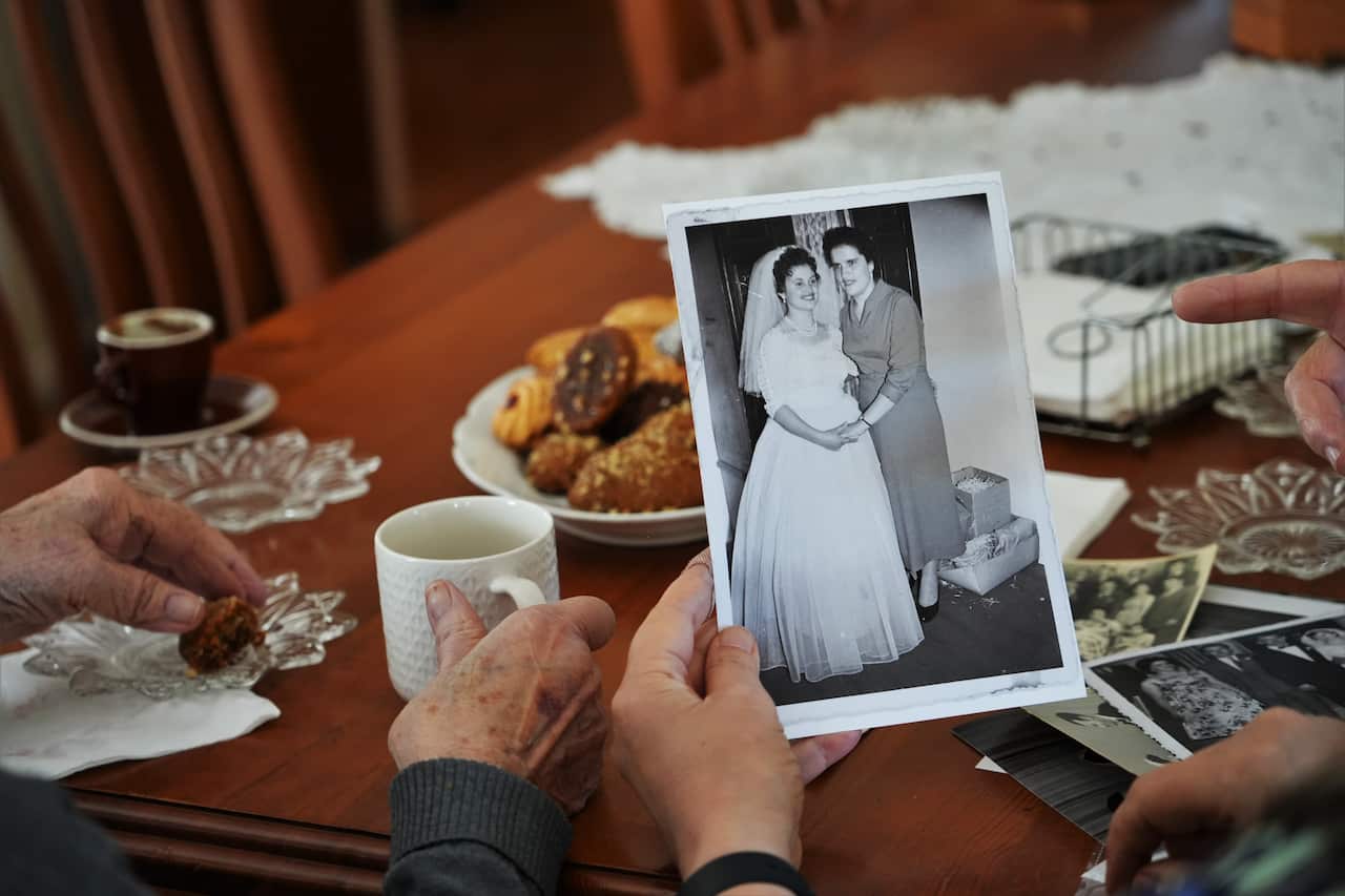 A person sits at a table holding a black-and-white photo of a woman standing next to a bride, with another pair of hands beside them holding a mug.