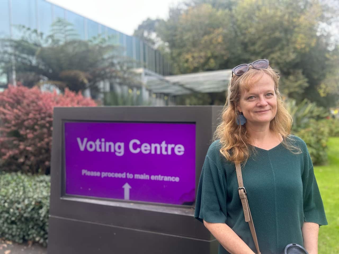 A woman wearing a green top stands next to a purple "voting centre" sign.