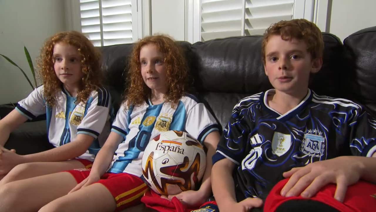 Three children with reddish hair are sitting together on a black leather couch wearing Argentina soccer jerseys, with the middle child holding a soccer ball.