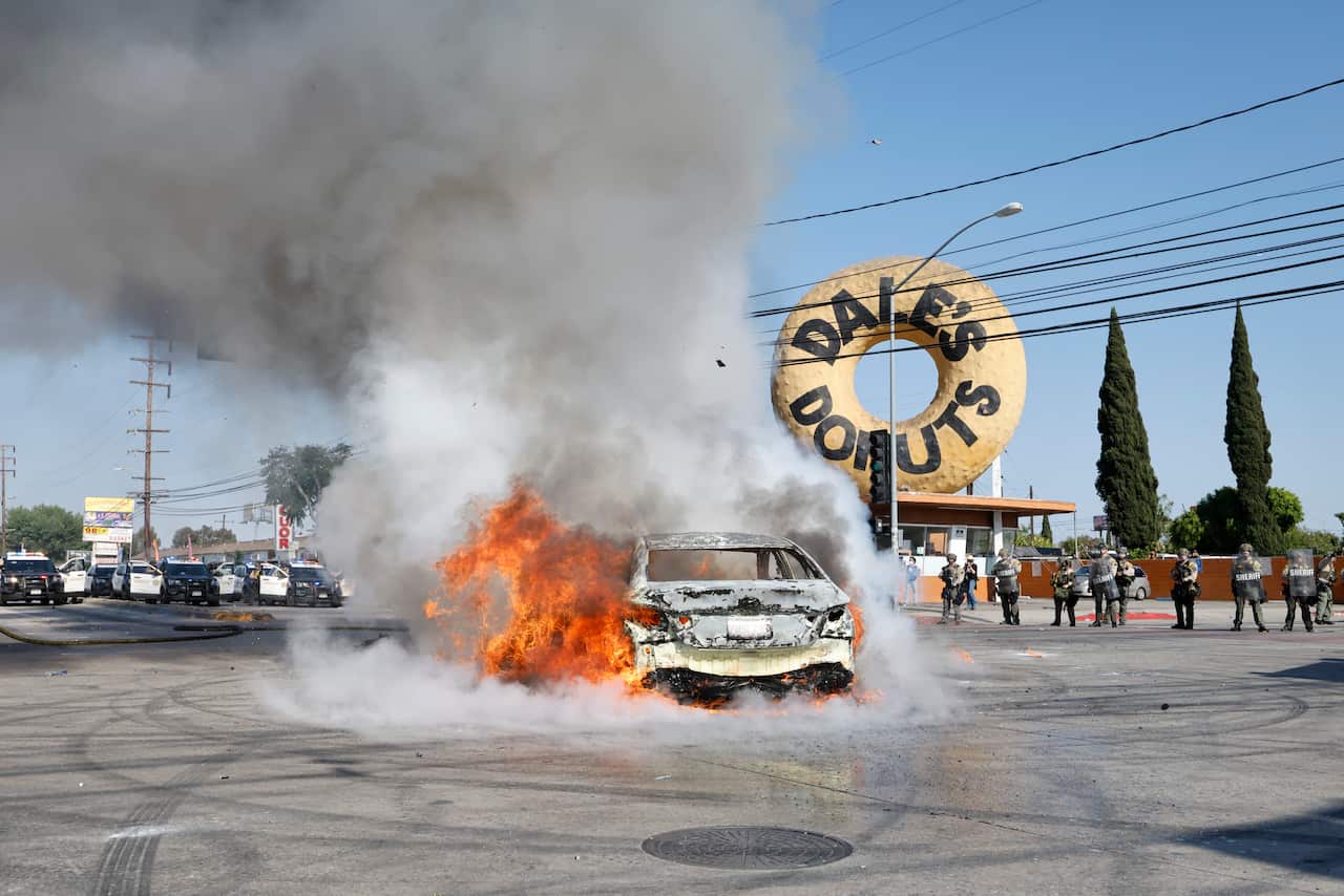 A vehicle on fire in the middle of the road, with police cars and officers standing in the distance, along with a donut shop.