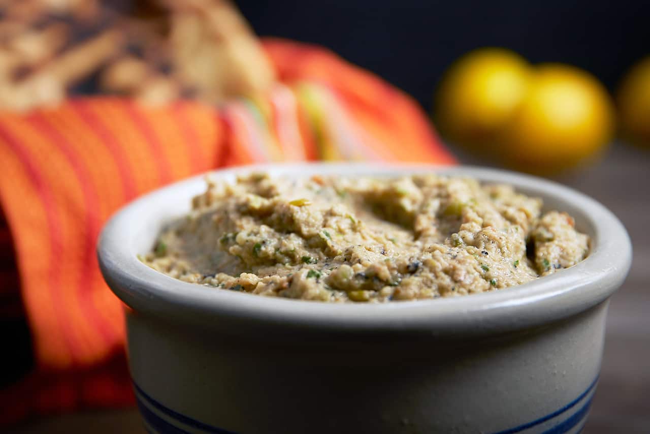 A greenish dip sits in a white ceramic pot. A basket, lined with a bright reddish towel and holding flatbreads, can be seen, out of focus, behind.  