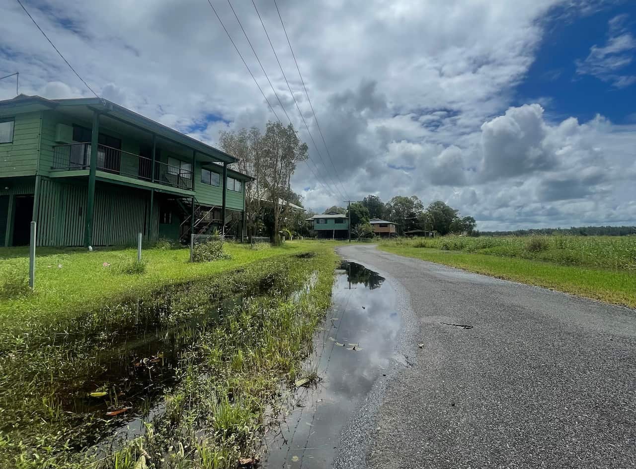 Puddle and wet grass in the foreground of a wide expanse of land with a green house on the left. Other homes are sparsely located in the background. 