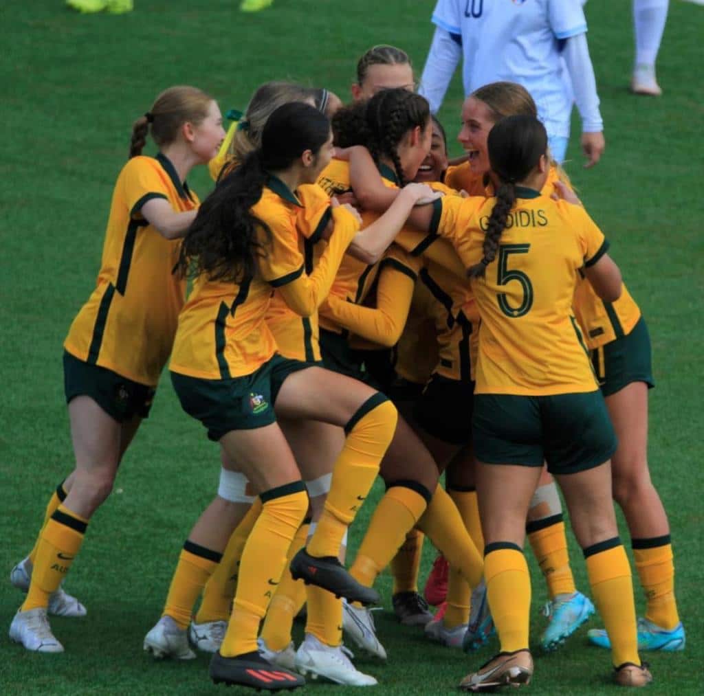 A group of young women in yellow football jerseys celebrating on a football pitch.