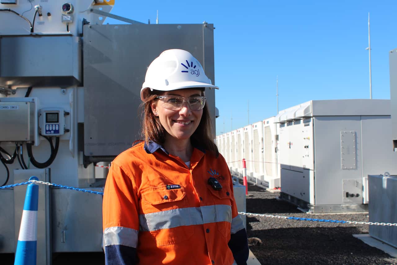 A woman wearing hi-vis, white hardhat and goggles stands outside. There is a long row of white cabinets stretching behind her