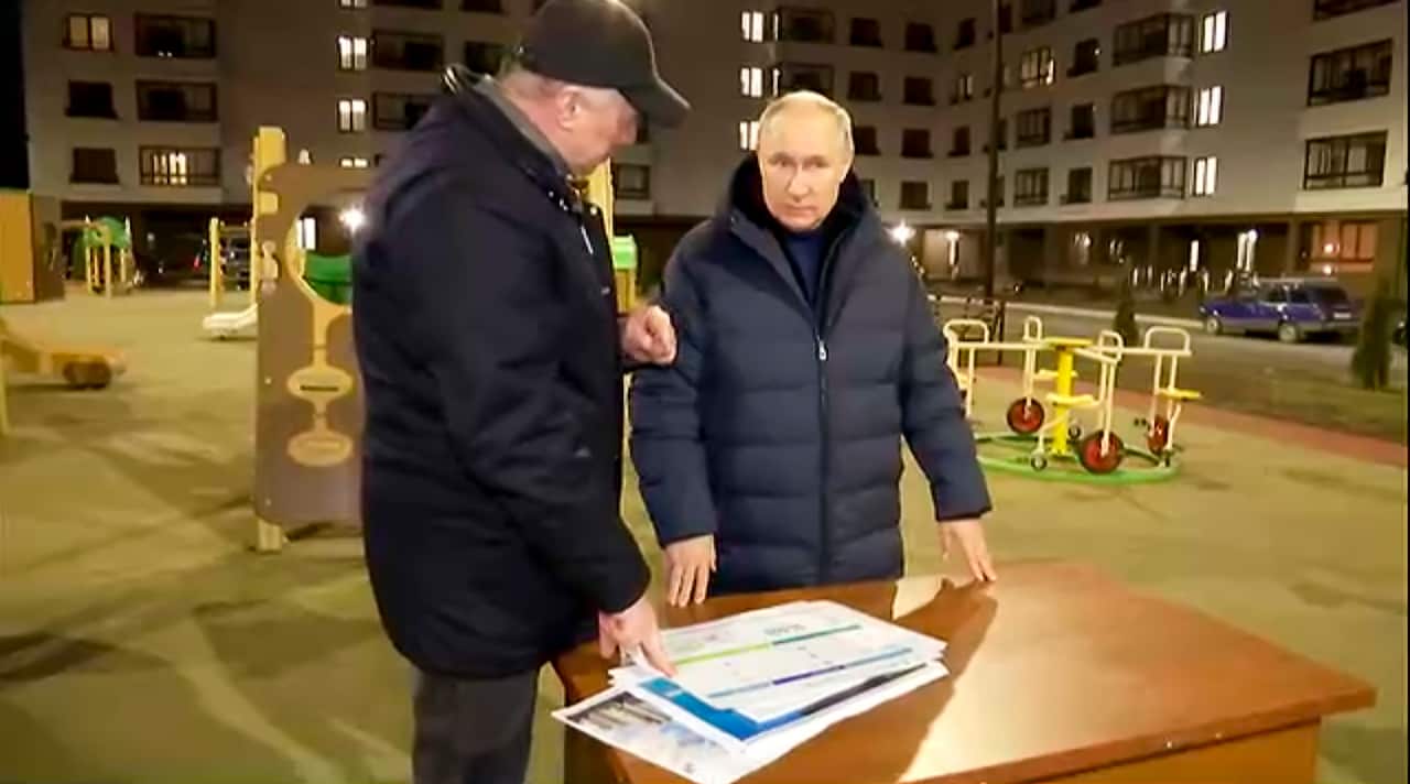 Putin and deputy stand at a desk near a kids park discussing papers. 