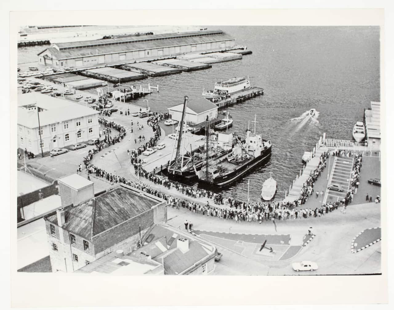 A black and white aerial photo showing people lining up along a harbour