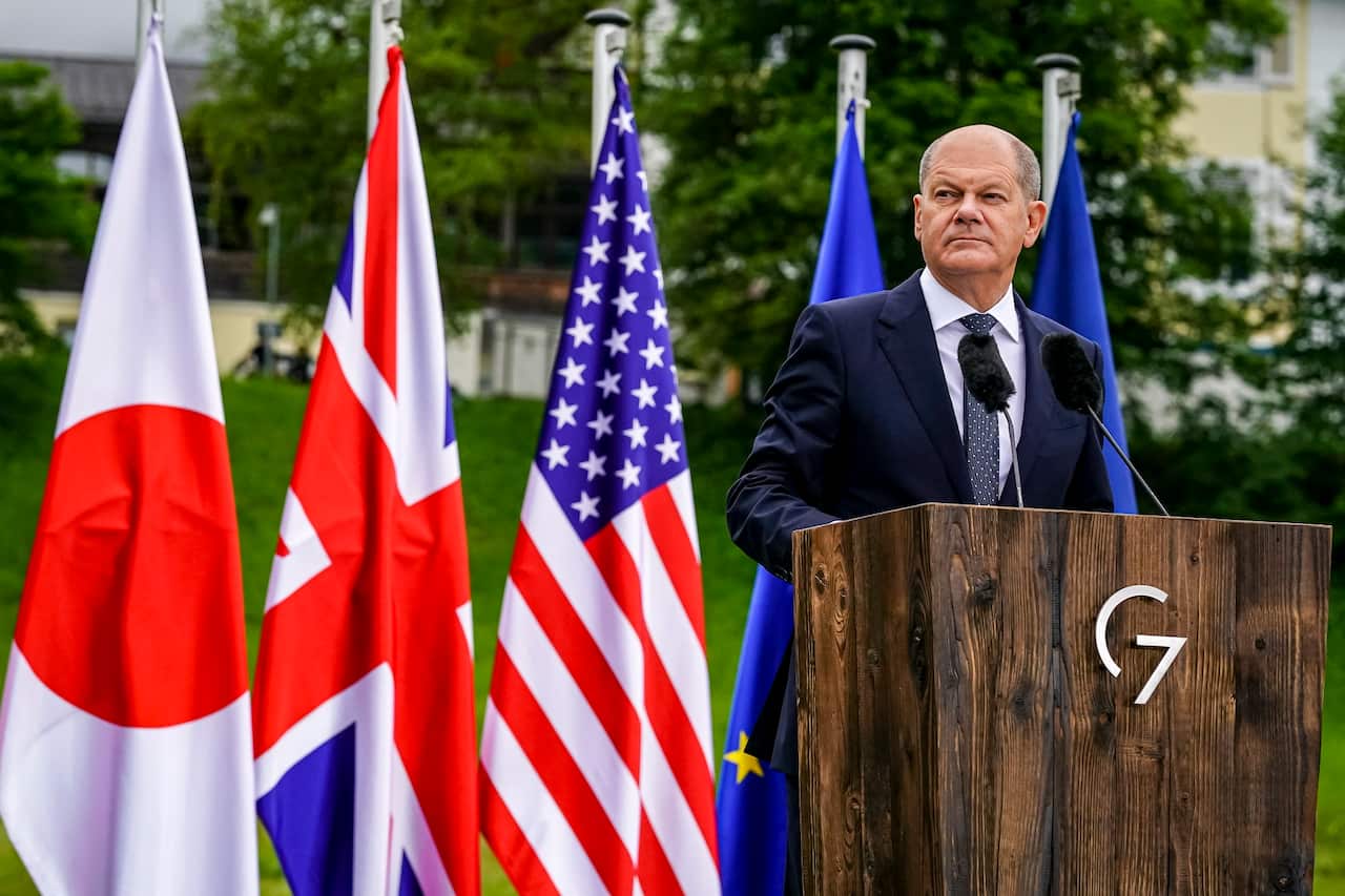German Chancellor and summit host Olaf Scholz stands at a podium with G7 member nation flags behind him.