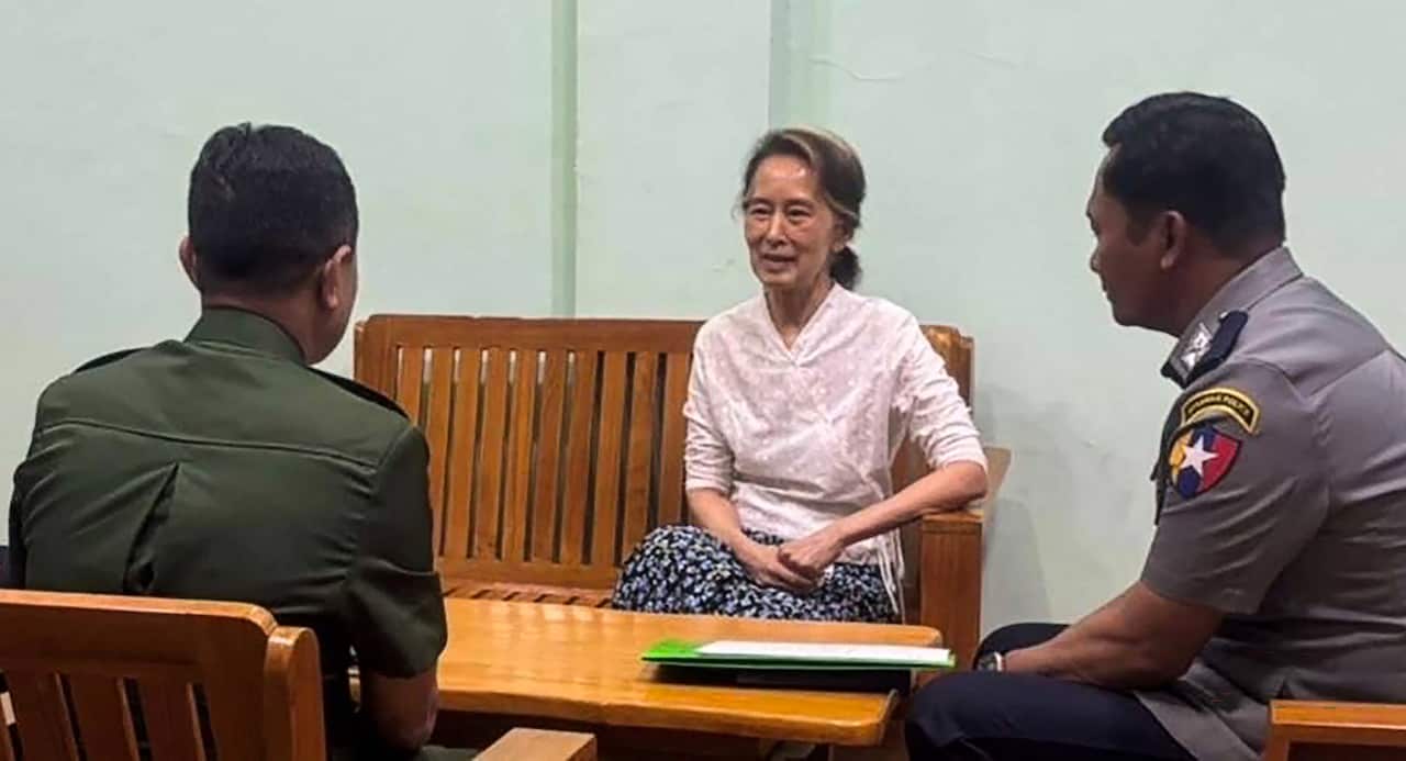 Aung San Suu Kyi sits on a wooden bench, flanked by two men in military attire.