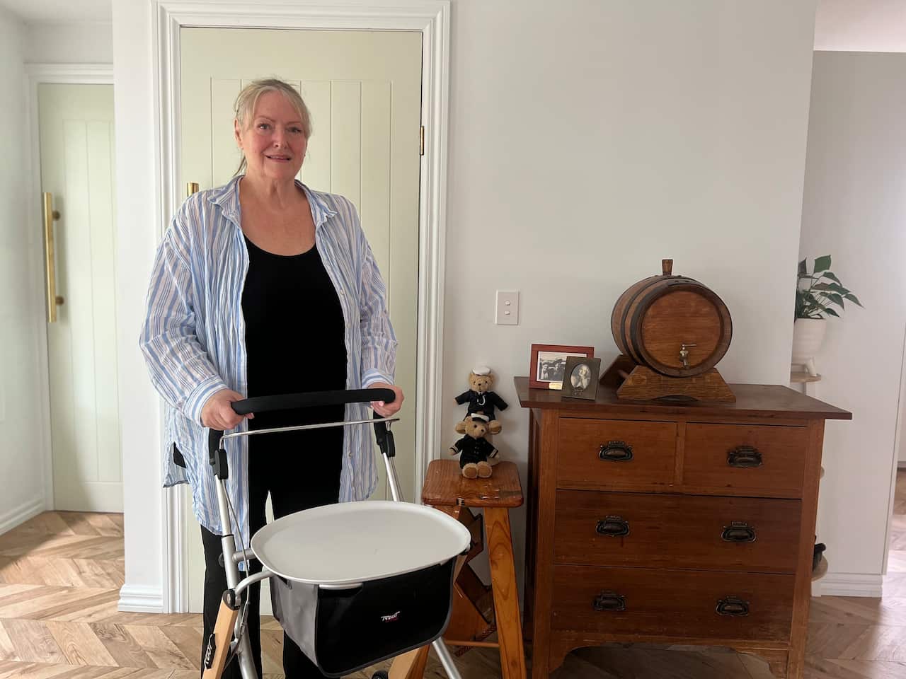 A woman with grey hair and wearing a black shirt and pants with a pale blue overshirt standing in front of a walker inside