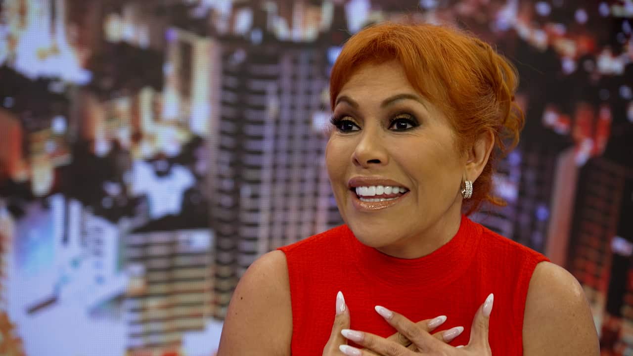 A glamorous woman with orange hair smiles, mid-sentence. Behind her is a background of a city. 