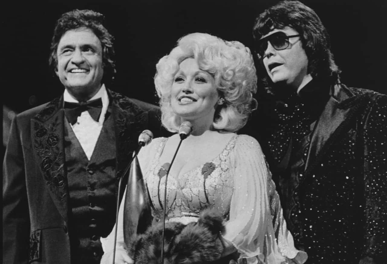 In a black-and-white photo, Dolly Parton speaks into a microphone. Johnny Cash and another man are standing next to her.