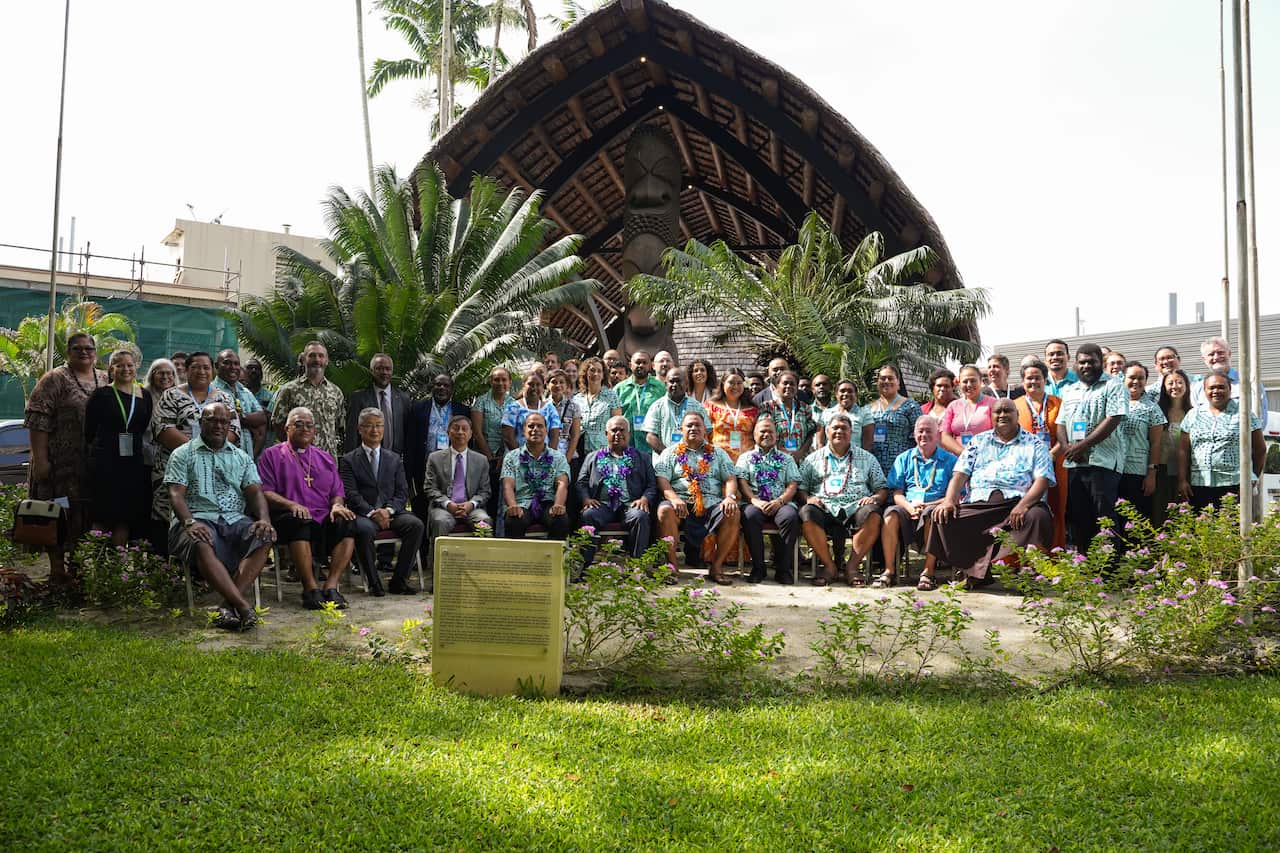 Approximately 35 people in bright shirts sit on three tears of chairs with a tropical backdrop