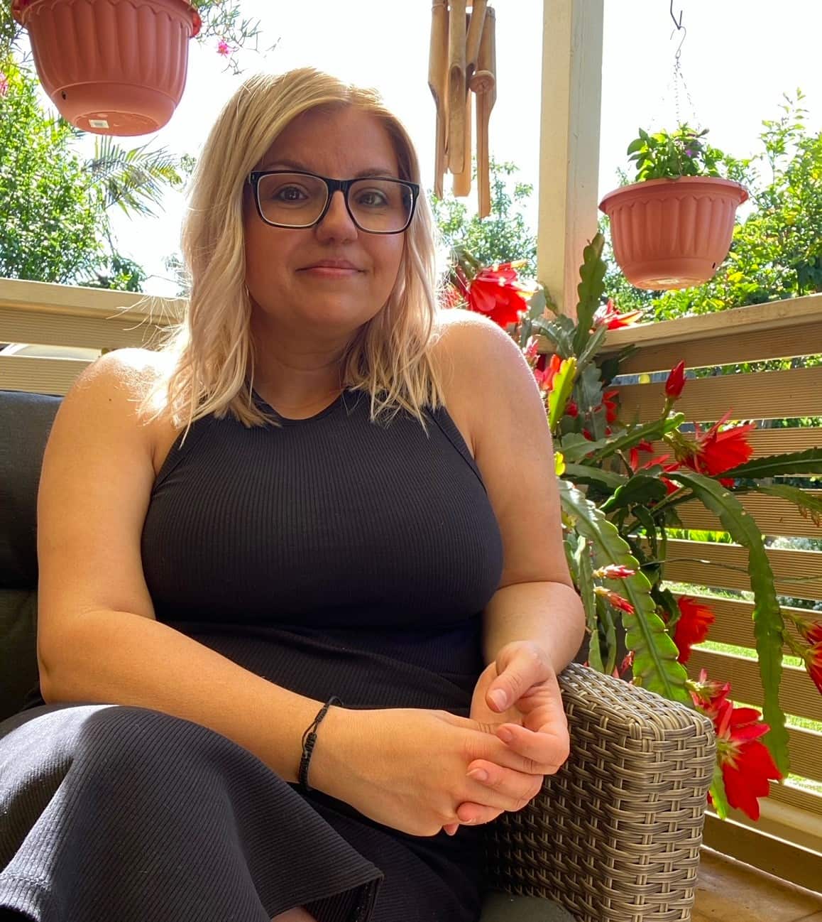A woman wearing a long black sleeveless dress sits on a balcony in a wicker chair