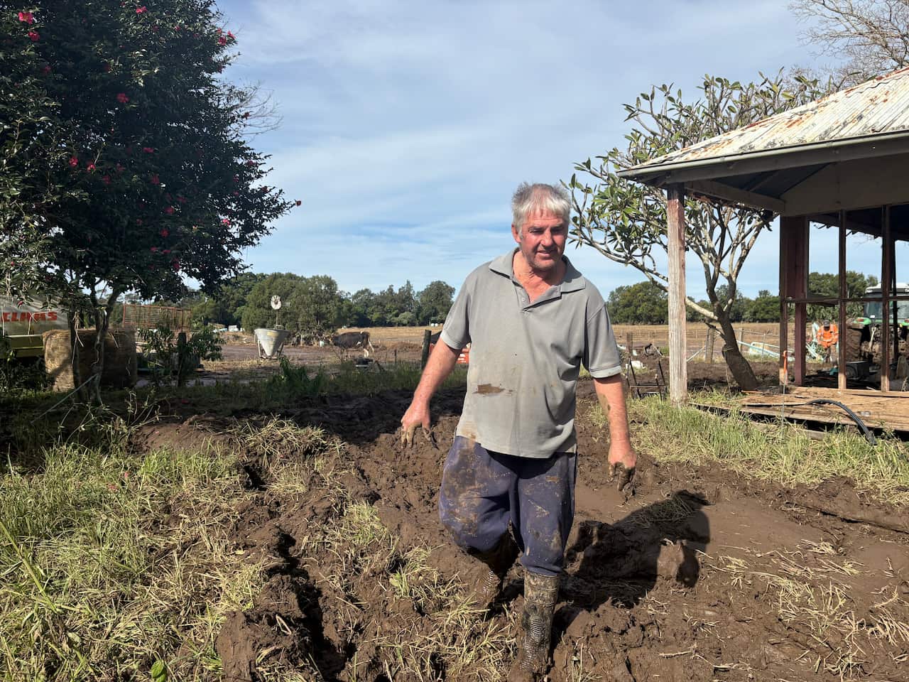 A middle aged man, slightly larger set, wearing a greyish shirt, and dark navy pants, both splattered with mud. He is walking toward the camera through thick mud. Behind him is a part of an old rundown house, a scene of farmland and a couple of cows.