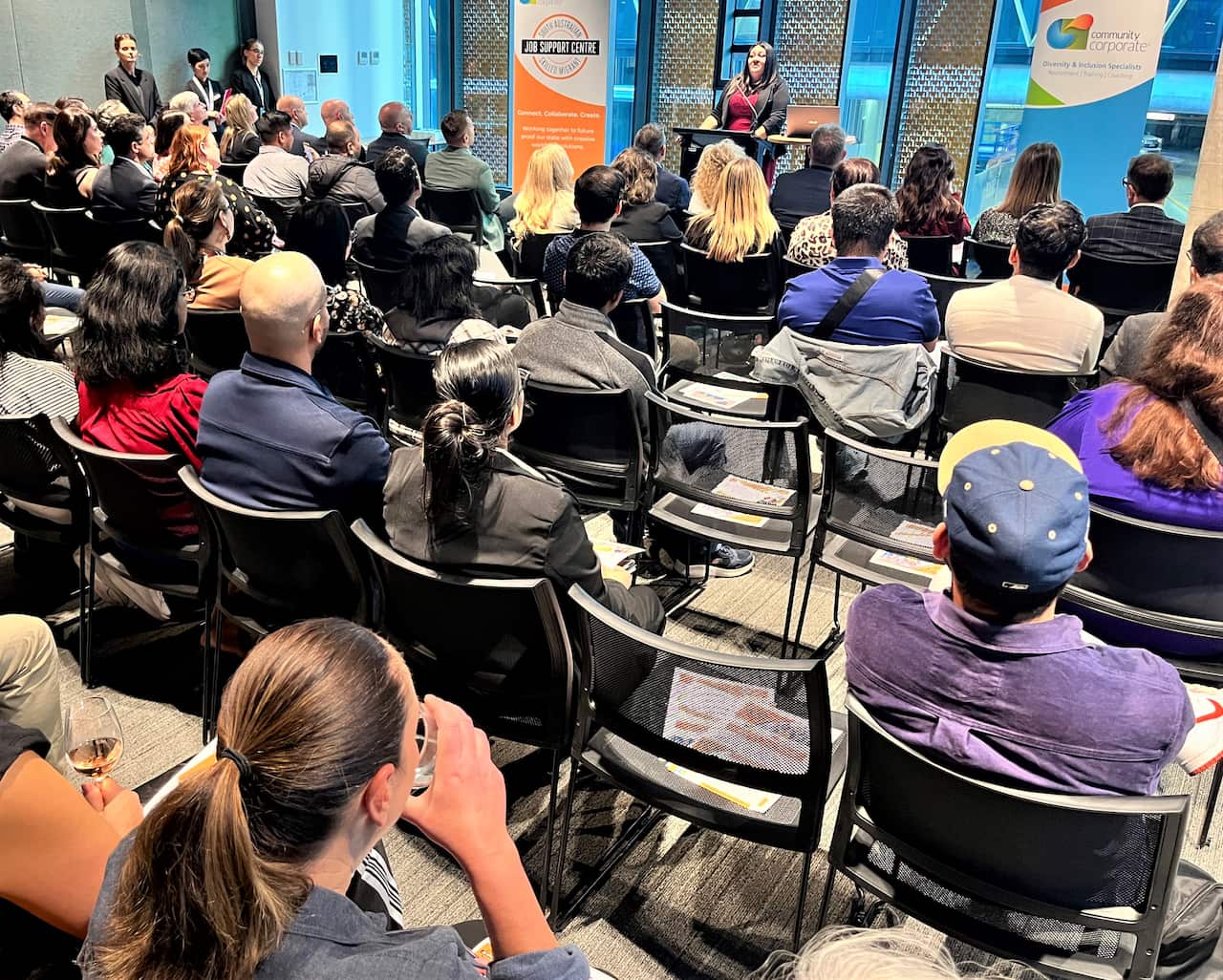A woman stands at a podium addressing a large group of people seated in rows of chairs.