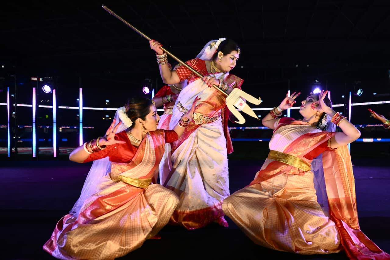 Dancers in golden and red saris are performing on a street. One holds a trident menacingly up to another.
