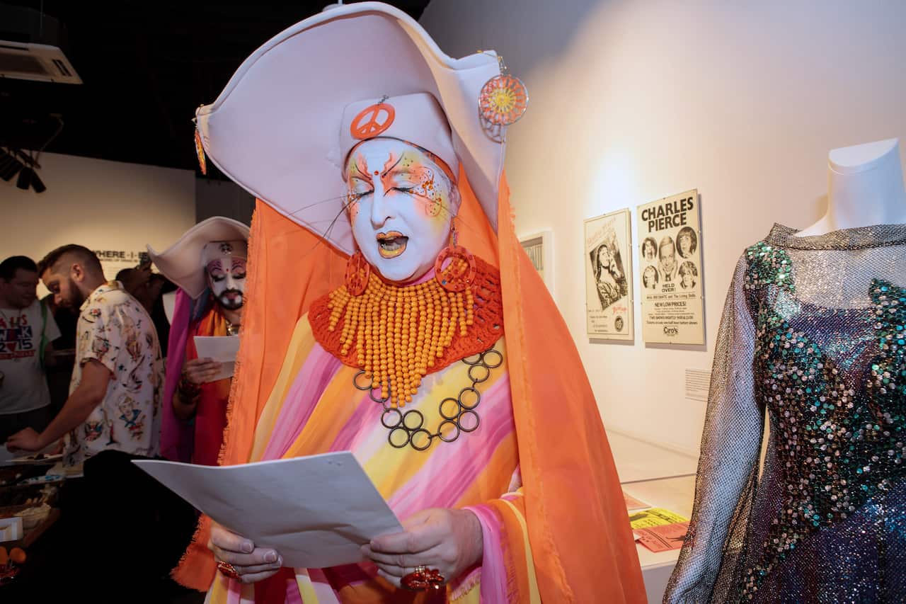 A drag queen, dressed in a pink, yellow, and orange nun costume, is reading from a document.