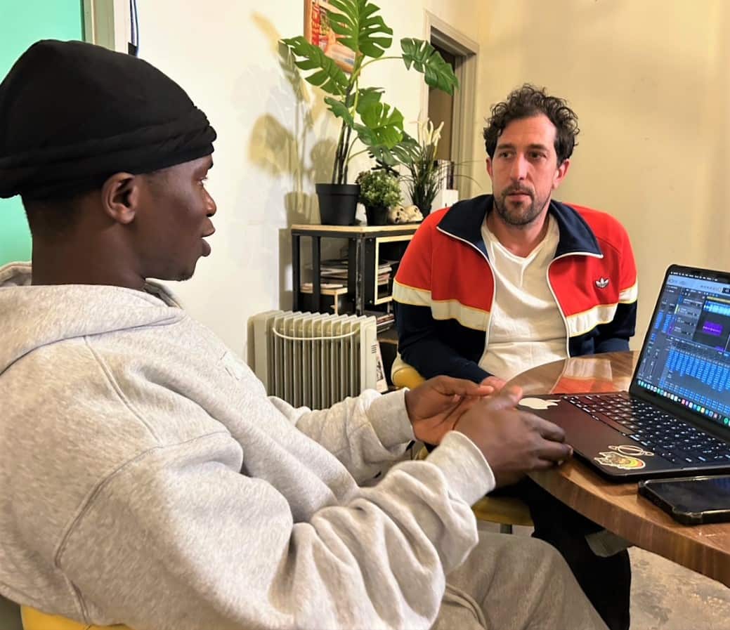 A man in a grey hoodie sits at a table with another man in a blue and red jacket.