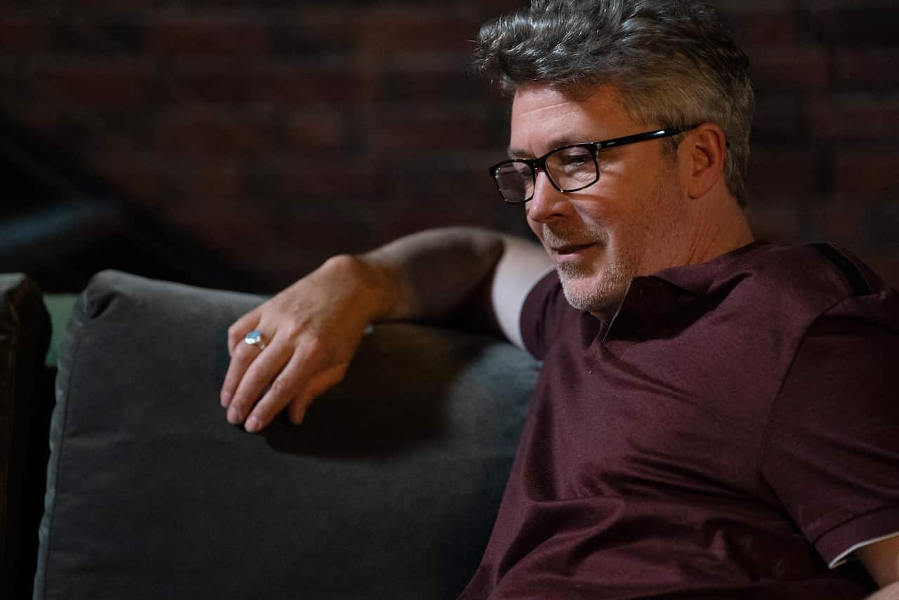 A man with stubble and glasses, in a maroom short-sleeved shirt, sits on a couch. He is smiling. 